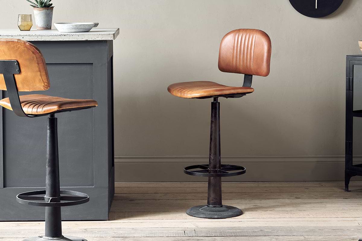The Narwana Bar Chair - Aged Tan by nkuku, featuring a brown leather seat and backrest, black metal base, and circular footrest, stands on a wooden floor next to a dark gray kitchen island with a light countertop. Another chair is partially visible.