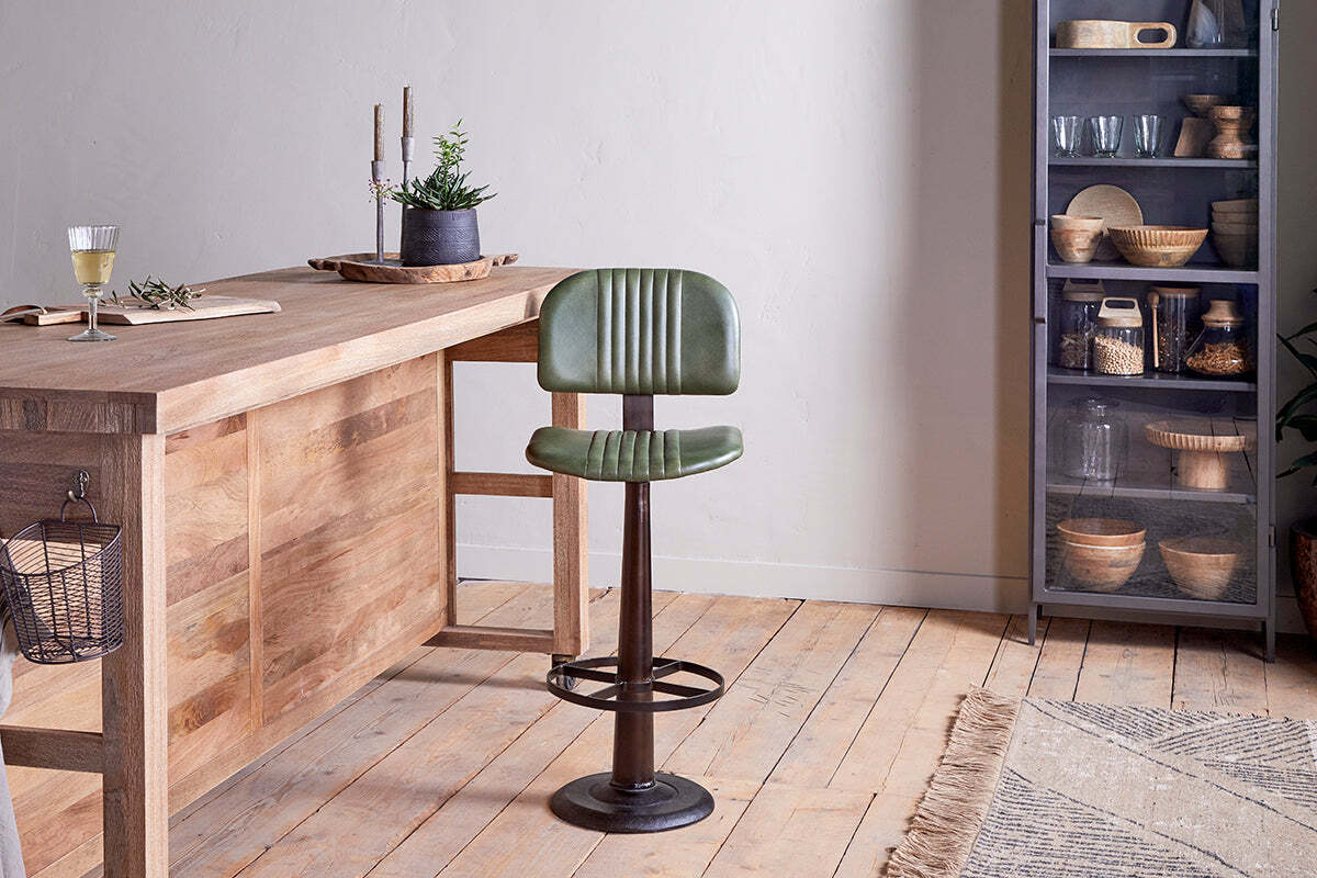 The Narwana Bar Chair - Rich Green by nkuku, a vintage-style swivel leather seat with backrest, stands on a wooden floor next to a wooden table with a drink and plant. A glass-front cabinet with bowls and glasses and a patterned rug complete the scene.