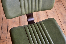 Two Narwana Bar Chairs in rich green by nkuku are shown from above. A dark metal support connects the industrial-style seats, which rest on a wooden floor for a modern bar stool effect.