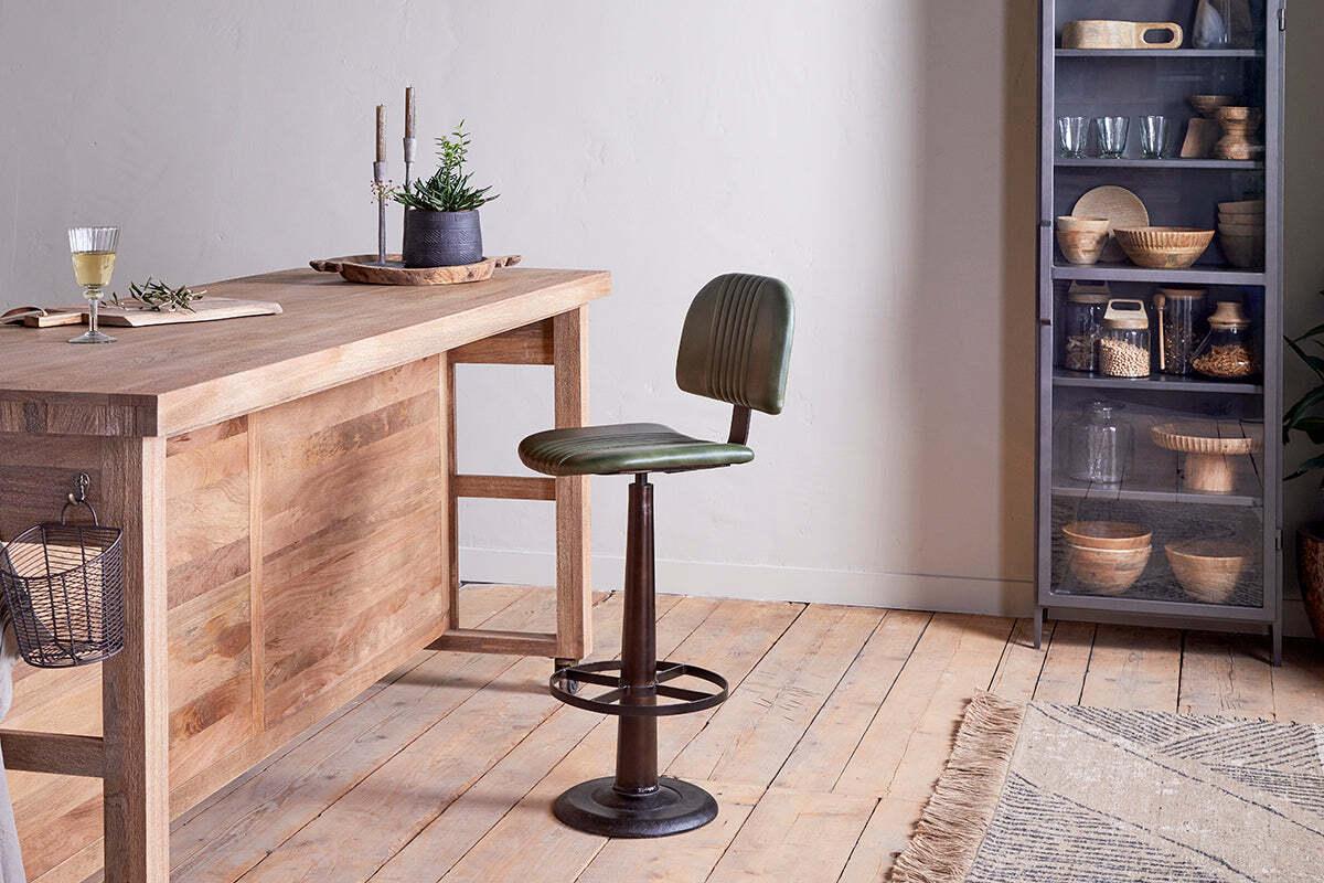 An industrial-style kitchen featuring a wooden island, the Narwana Bar Chair - Rich Green by nkuku, and a glass-door cabinet displaying kitchenware.