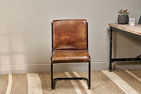 The Narwana Leather Desk Chair - Aged Tan by nkuku features a brown leather seat and backrest on a sleek iron frame. It stands on a beige woven rug beside a wooden desk with black metal legs and two small potted plants.