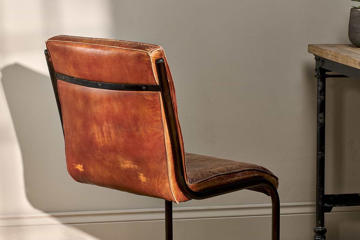 The Narwana Leather Desk Chair - Aged Tan by nkuku sits beside a wooden table with a black metal frame, sunlight highlighting the chair’s aged leather texture and iron backrest.