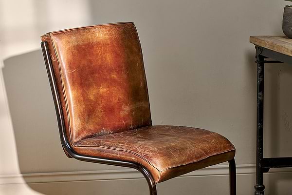 A close-up of the Narwana Leather Desk Chair - Aged Tan by nkuku showcases its vintage-style distressed brown leather, curved iron frame, and padded seat and backrest—an ideal piece to elevate any interior space.