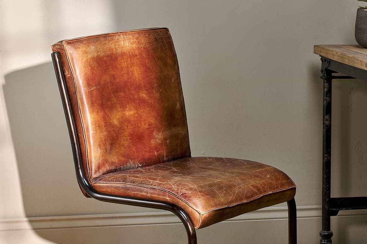 A close-up of the Narwana Leather Desk Chair - Aged Tan by nkuku showcases its vintage-style distressed brown leather, curved iron frame, and padded seat and backrest—an ideal piece to elevate any interior space.