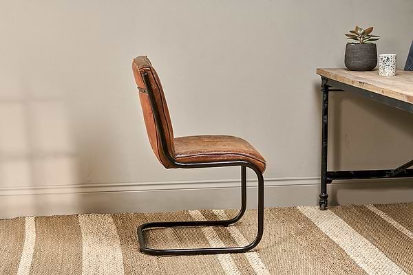 The Narwana Leather Desk Chair in aged tan by nkuku, with an iron frame, sits on a beige woven rug beside a wooden desk with black metal legs. The desk holds a small potted succulent and white patterned cup, set against a light gray wall for a calm look.