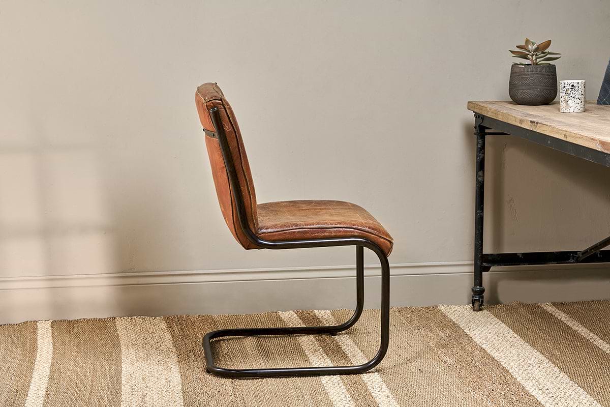 The Narwana Leather Desk Chair in aged tan by nkuku, with an iron frame, sits on a beige woven rug beside a wooden desk with black metal legs. The desk holds a small potted succulent and white patterned cup, set against a light gray wall for a calm look.