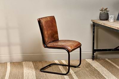 The Narwana Leather Desk Chair in Aged Tan by nkuku, featuring an iron frame, is placed on a beige striped rug next to a wooden desk with a succulent and patterned cup, lending warmth to the inviting space against a light wall.