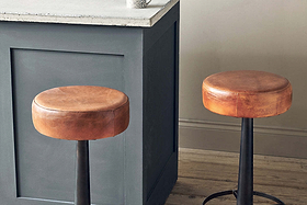 Two Nkuku Narwana Leather Round Stools in aged tan with black metal bases are set by a gray paneled kitchen island with a white countertop, on light wood flooring beside a beige wall.