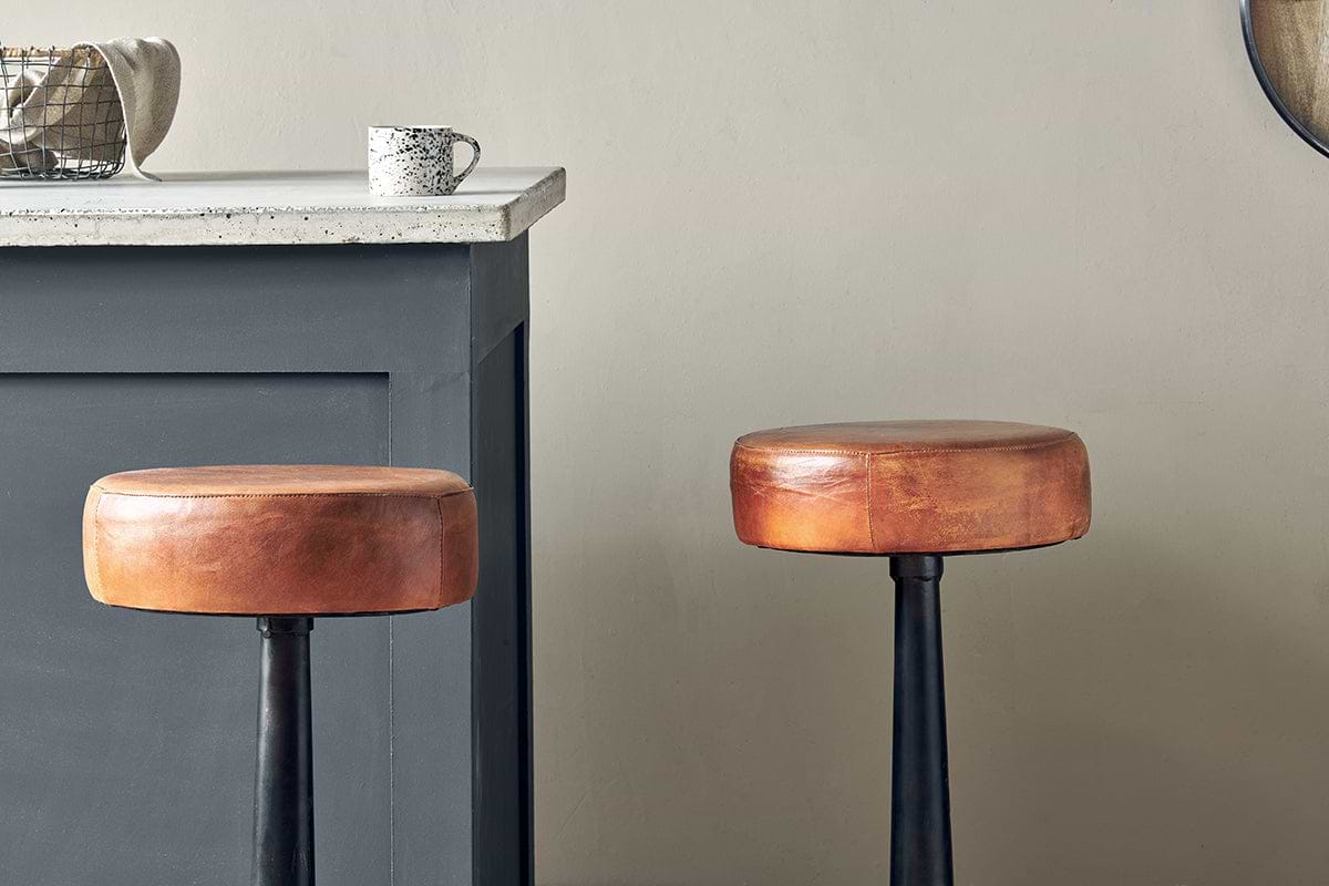 Two nkuku Narwana Leather Round Stools in aged tan, each with round seats and black metal legs, stand by a gray kitchen counter topped with a speckled mug and small basket. The background wall is light beige.
