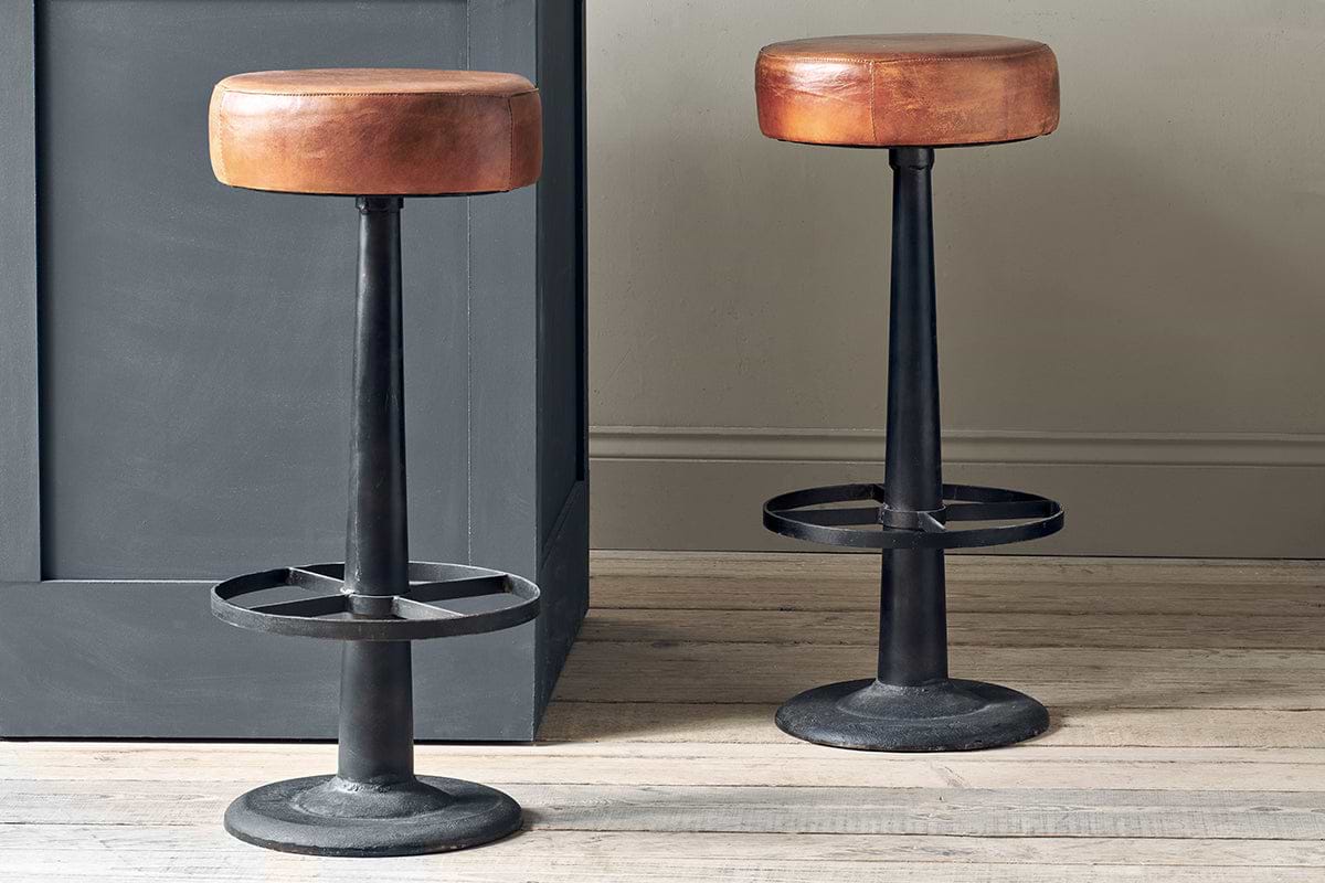 Two nkuku Narwana Leather Round Stools in Aged Tan, featuring round leather seats, black metal bases with footrests, and a sturdy minimalist design, stand on a light wooden floor by a gray paneled counter.