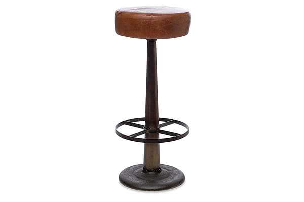The Narwana Leather Round Stool - Aged Tan by nkuku features an aged tan leather seat, single dark metal pedestal base, and circular footrest. This counter stool has an industrial, minimalist design set against a white background.