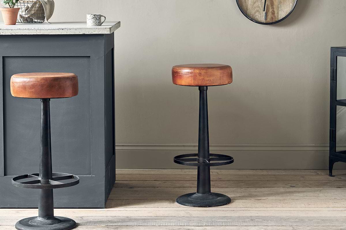 Two nkuku Narwana Leather Round Stools in aged tan, featuring round seats and black metal bases, stand on a light wooden floor by a gray kitchen island. A potted plant and mug sit on the counter, with a clock on the neutral wall behind.
