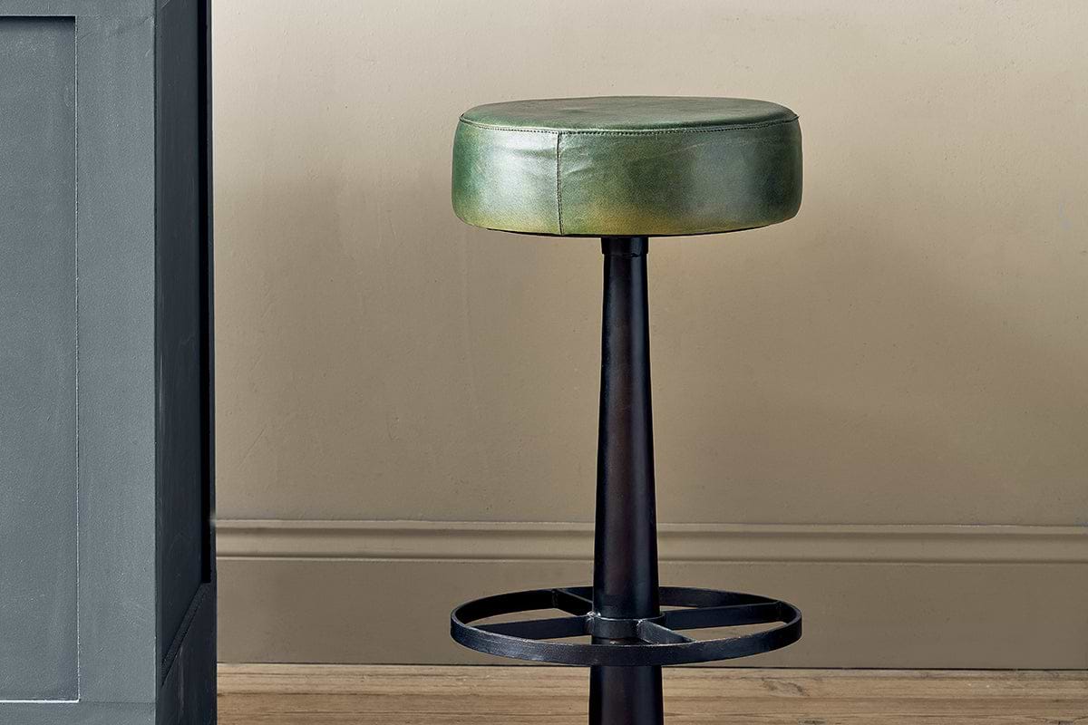 The Narwana Leather Round Stool by nkuku features a rich green cushioned seat, black metal base, and circular footrest. Ideal for a kitchen island, it stands on wood flooring beside a gray counter against a beige wall.