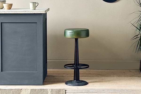 The nkuku Narwana Leather Round Stool in rich green, featuring a black metal base and circular footrest, stands on a light wood floor by a gray kitchen island with bowls, a mug, and a plant nearby against neutral walls.