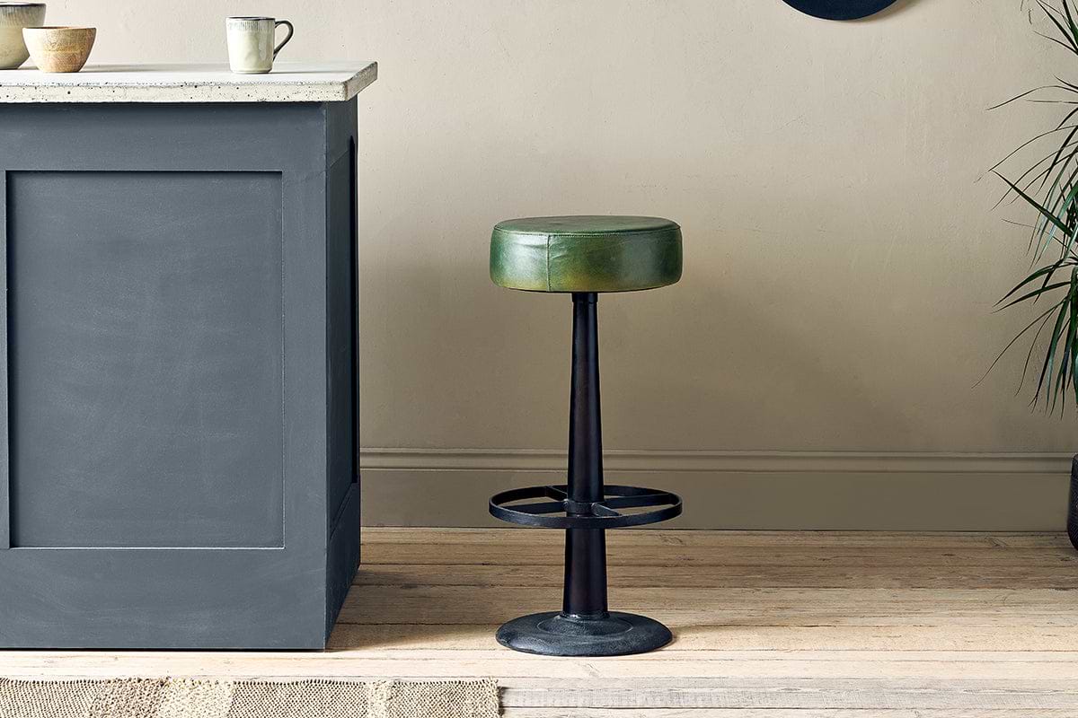 The nkuku Narwana Leather Round Stool in rich green, featuring a black metal base and circular footrest, stands on a light wood floor by a gray kitchen island with bowls, a mug, and a plant nearby against neutral walls.