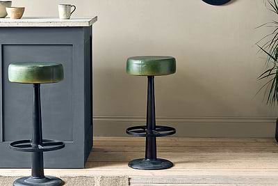 Two nkuku Narwana Leather Round Stools in Rich Green, featuring black metal bases and footrests, are placed beside a dark grey kitchen island.