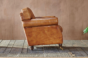 The nkuku Narwana Ribbed Leather Armchair in Aged Tan is showcased in profile on a rustic wooden floor, set against a textured brown wall with a leafy green plant partially visible to the right.