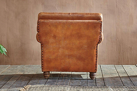 Back view of the Narwana Ribbed Leather Armchair - Aged Tan by nkuku, featuring ribbed stitching, nailhead trim, and turned wooden legs on rustic floorboards, with a textured rug and green plant at the edges against a beige wall.