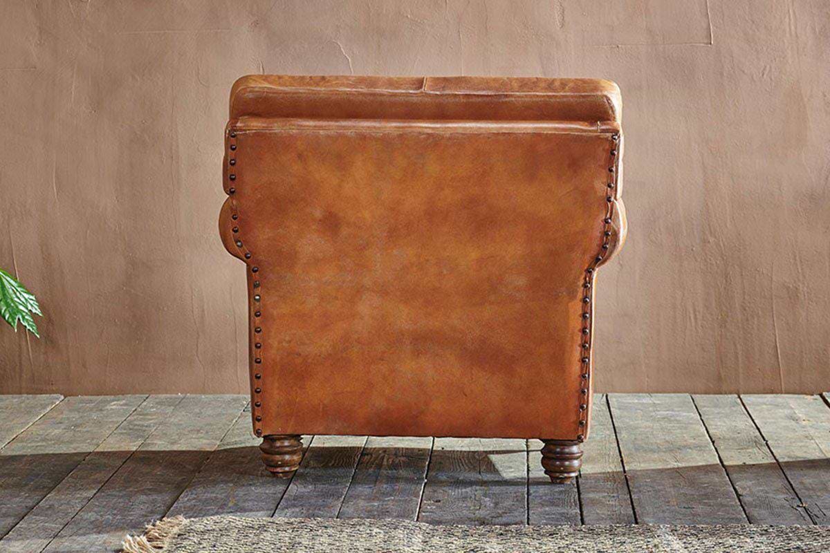 Back view of the Narwana Ribbed Leather Armchair - Aged Tan by nkuku, featuring ribbed stitching, nailhead trim, and turned wooden legs on rustic floorboards, with a textured rug and green plant at the edges against a beige wall.