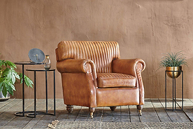 An nkuku Narwana Ribbed Leather Armchair in aged tan sits on a wooden floor, flanked by two black metal side tables with a clock and glass on the left, and a gold planter with green foliage to the right, against a plain brown wall.