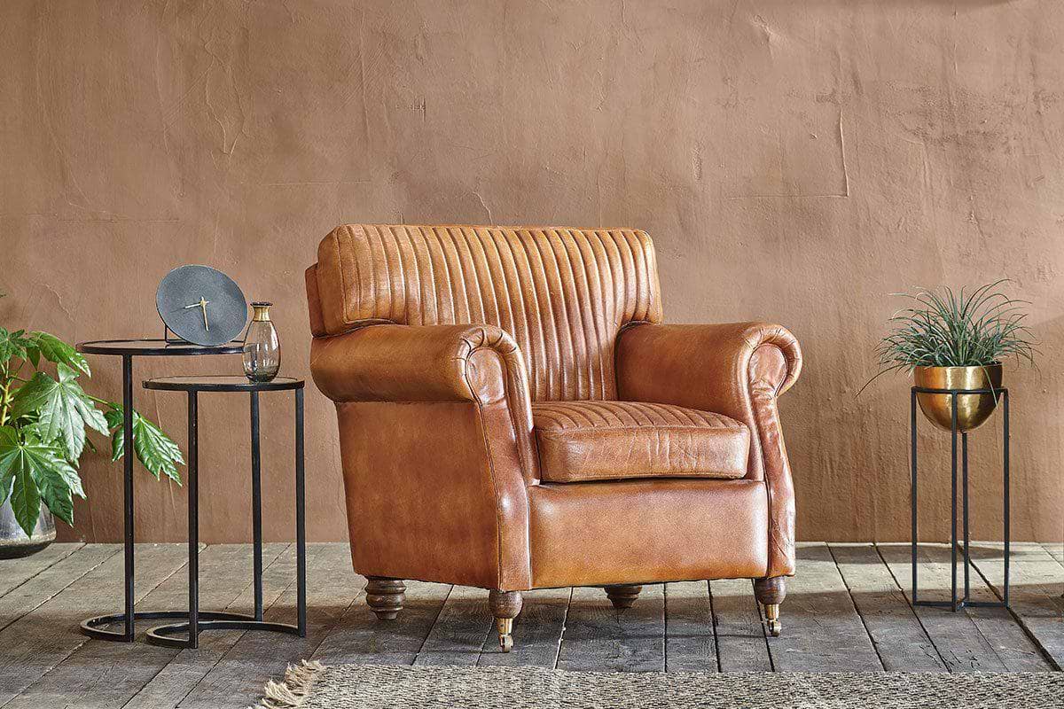 An nkuku Narwana Ribbed Leather Armchair in aged tan sits on a wooden floor, flanked by two black metal side tables with a clock and glass on the left, and a gold planter with green foliage to the right, against a plain brown wall.