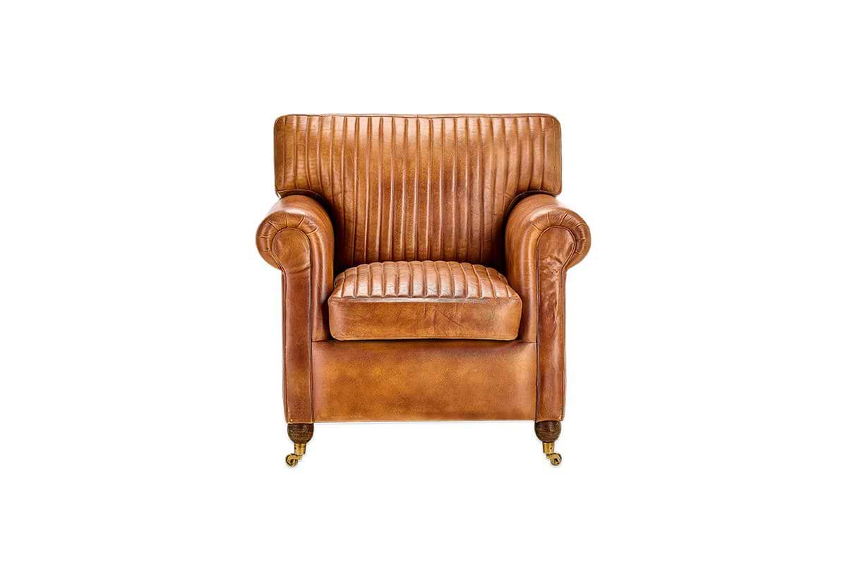 The nkuku Narwana Ribbed Leather Armchair - Aged Tan features ribbed stitching, rolled arms, and brass caster wheels on four legs, shown against a plain white background.