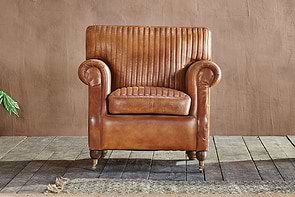 The Narwana Ribbed Leather Armchair - Aged Tan by nkuku, featuring ribbed stitching, rolled arms, and short wooden legs with brass casters, rests on a rustic floor beside a textured rug and leafy plant for an inviting touch.