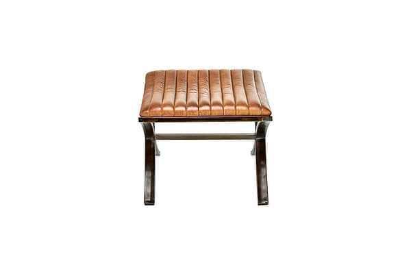 The Narwana Ribbed Leather Footstool – Aged Tan by nkuku features a brown ribbed leather cushion with vertical stitching and a dark metal X-shaped base, shown from the front against a plain white background.