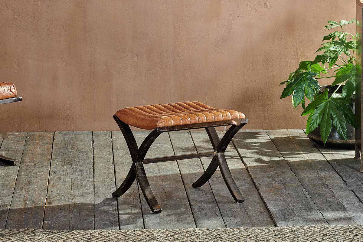 The Narwana Ribbed Leather Footstool in aged tan by nkuku features dark curved legs and sits on a rustic wooden floor. It's ideal for living rooms or bedrooms, paired with the matching chair and a green plant against a light brown wall.