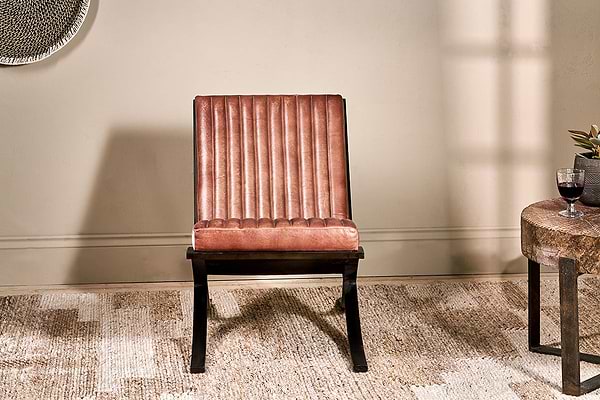 The nkuku Narwana Ribbed Leather Lounger in aged tan, featuring a padded seat, vertical stitching, and dark wood legs, sits on a beige rug. A round side table with a plant and red wine is nearby, with soft light casting shadows on the neutral wall.