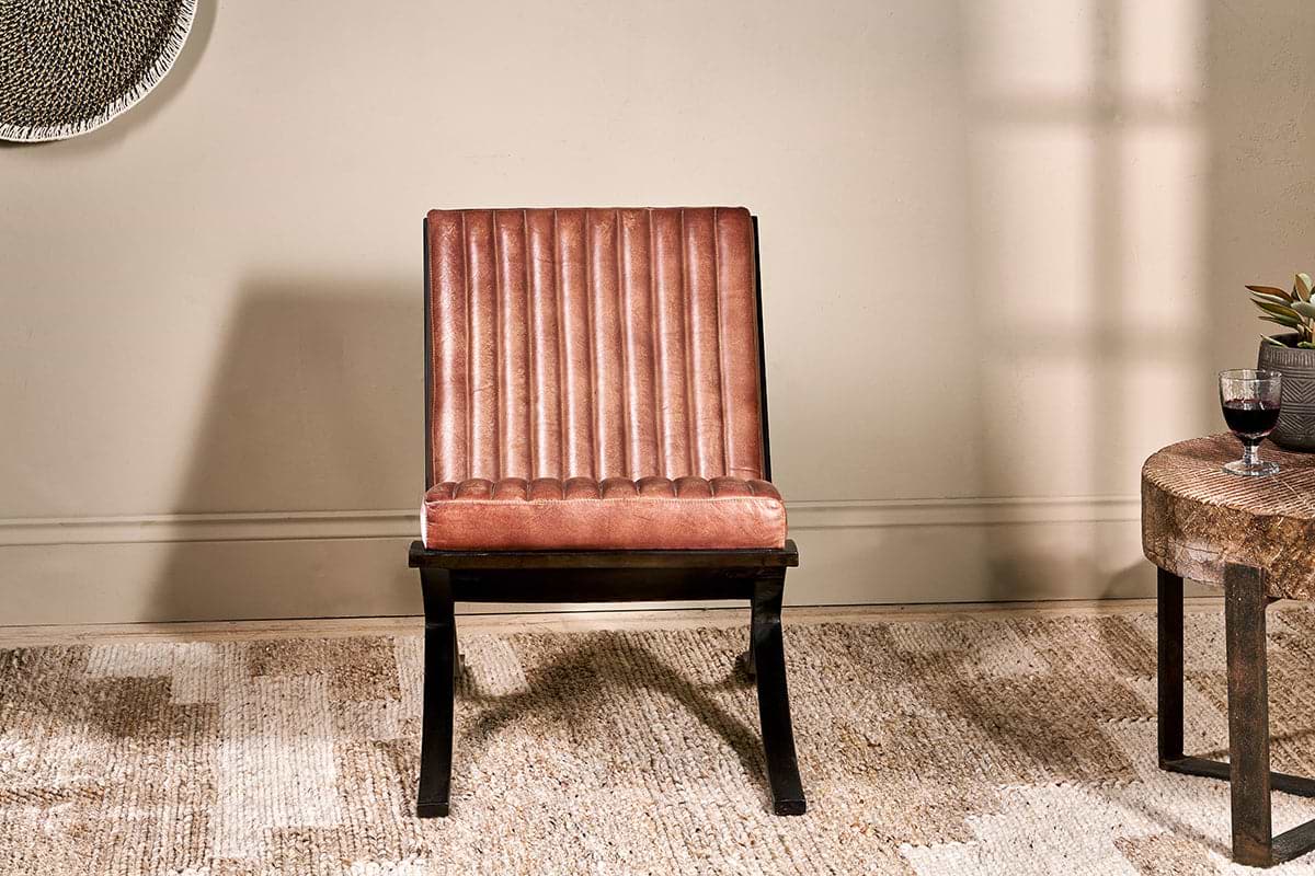 The nkuku Narwana Ribbed Leather Lounger in aged tan, featuring a padded seat, vertical stitching, and dark wood legs, sits on a beige rug. A round side table with a plant and red wine is nearby, with soft light casting shadows on the neutral wall.