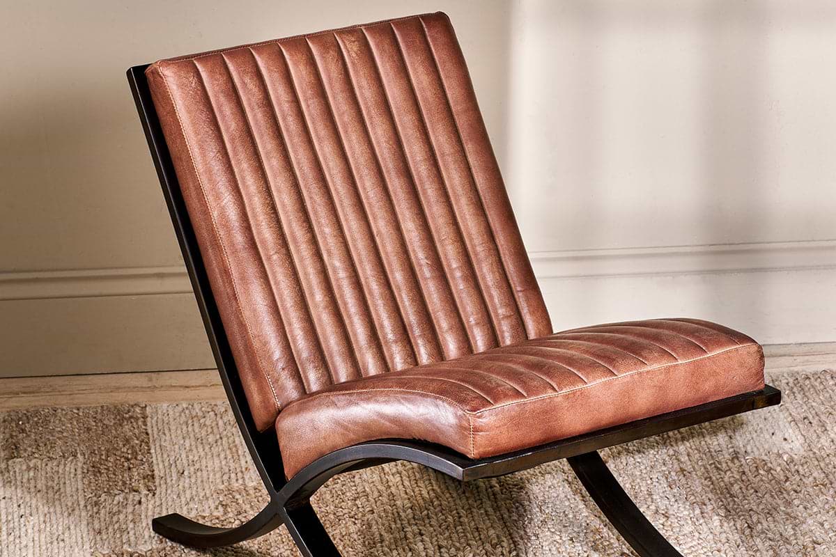 The Narwana Ribbed Leather Lounger - Aged Tan by nkuku features a padded seat with brown vertical ribbed stitching, black curved metal legs, and sits on a textured beige rug against a light-colored wall.