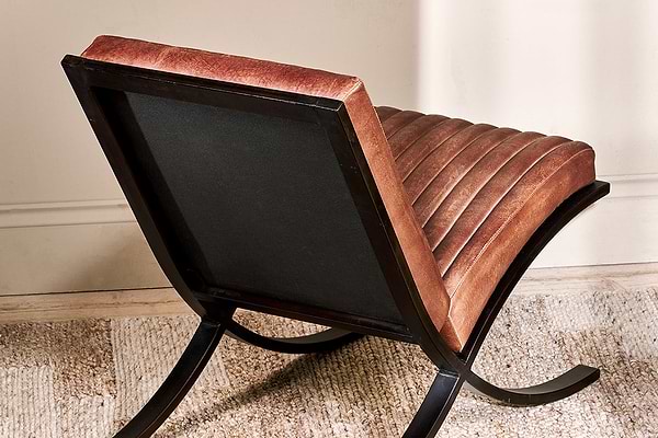 A close-up of the Narwana Ribbed Leather Lounger in Aged Tan by nkuku, featuring ribbed stitching, channel tufting, a padded seat, and a dark wood frame with curved legs.