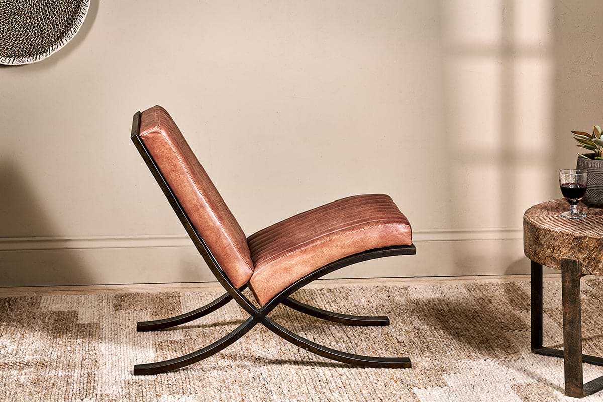 The Narwana Ribbed Leather Lounger by nkuku, with aged tan leather cushions and a curved black metal frame, sits on a beige textured rug beside a small table with red wine and a plant, sunlight casting soft shadows.