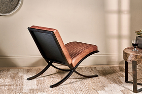 The nkuku Narwana Ribbed Leather Lounger - Aged Tan, featuring a brown ribbed leather seat and black metal frame, sits on a woven rug beside a wooden side table topped with a glass of red wine.