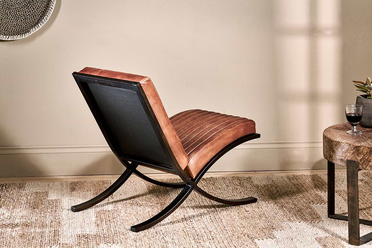 The nkuku Narwana Ribbed Leather Lounger - Aged Tan, featuring a brown ribbed leather seat and black metal frame, sits on a woven rug beside a wooden side table topped with a glass of red wine.