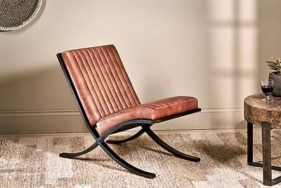 The nkuku Narwana Ribbed Leather Lounger in Aged Tan with padded seat rests on a woven rug. To the right, a rustic wooden side table holds a glass of red wine and a potted plant.
