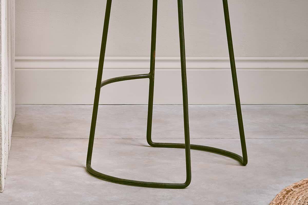 Close-up of the dark metal base and thin, curved legs with a support bar on the nkuku Narwana Ribbed Leather Stool - Aged Tan - Large, set against a textured, light-colored wall with molding in the background.