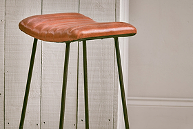 The Narwana Ribbed Leather Stool - Aged Tan (Large) by nkuku features a ribbed leather seat and slim black metal legs, adding a stylish mix of textures and tones to your minimal space against white paneling and beige wainscoting.