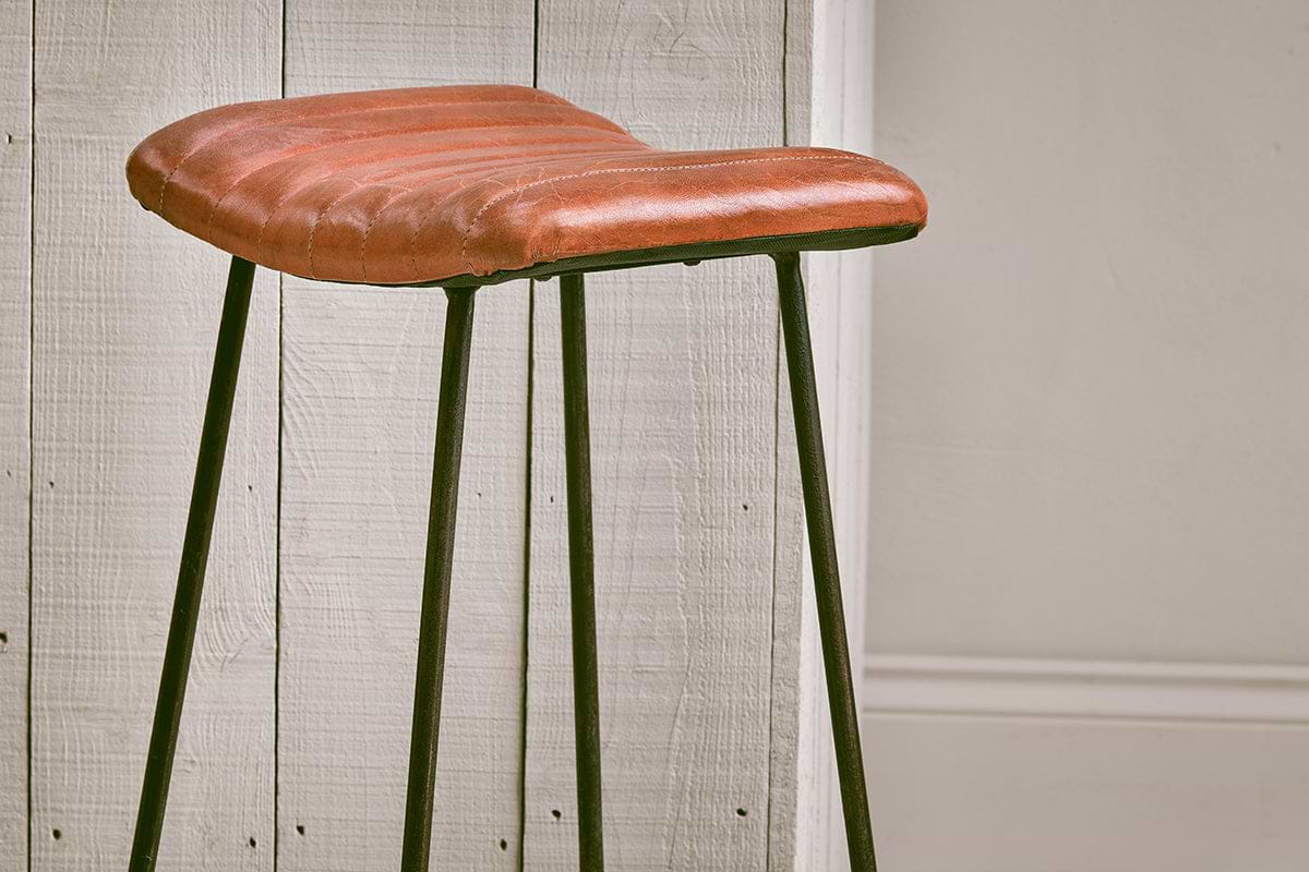 The Narwana Ribbed Leather Stool - Aged Tan (Large) by nkuku features a ribbed leather seat and slim black metal legs, adding a stylish mix of textures and tones to your minimal space against white paneling and beige wainscoting.