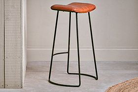 The Narwana Ribbed Leather Stool in Aged Tan (Large) by nkuku, featuring a padded seat and dark metal base, sits in front of a pale gray wall.
