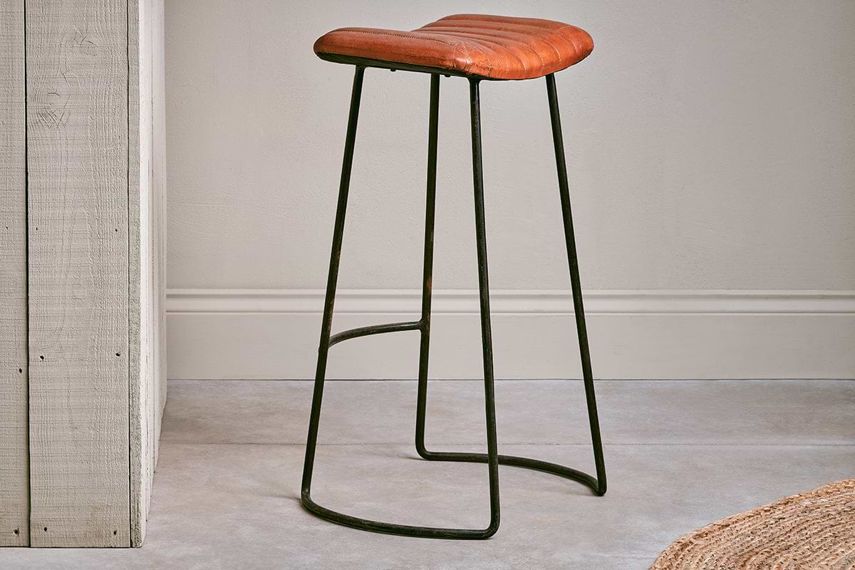 The Narwana Ribbed Leather Stool in Aged Tan (Large) by nkuku, featuring a padded seat and dark metal base, sits in front of a pale gray wall.