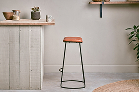 A minimalist kitchen with a wooden countertop, small potted plants, and jars on shelves. The nkuku Narwana Ribbed Leather Stool in Aged Tan (Large) with black metal legs sits on the gray floor. A jute rug and leafy plant are partially visible in front.