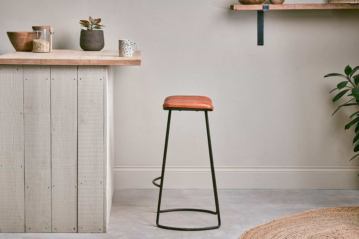 A minimalist kitchen with a wooden countertop, small potted plants, and jars on shelves. The nkuku Narwana Ribbed Leather Stool in Aged Tan (Large) with black metal legs sits on the gray floor. A jute rug and leafy plant are partially visible in front.
