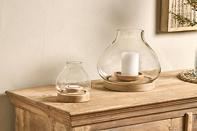 Two nkuku Naryla Mango Wood Lanterns made of recycled glass and natural mango wood sit on a rustic cabinet—one with a pillar candle, one with a tealight. A framed textured wall art is partially visible in the upper right corner.