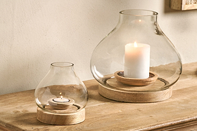 Two nkuku Naryla Mango Wood Lanterns in natural finish, one with a tealight and the other with a pillar candle, cast warm light from their mango wood bases and recycled glass tops on a wooden surface against a plain wall.