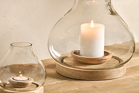 Two nkuku Naryla Mango Wood Lanterns - Natural, featuring mango wood bases and recycled glass, sit on a light wooden surface—one with a white pillar candle, the other with a tealight—casting a warm, soft glow.