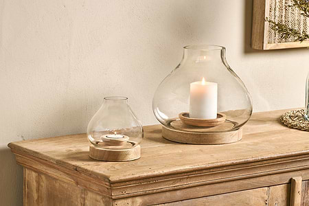 Two nkuku Naryla Mango Wood Lanterns in natural, each with a recycled glass top and mango wood base, sit on a rustic cabinet; the larger holds a pillar candle, the smaller cradles a tealight against a neutral-toned wall.