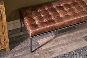 The Nasan Leather Upholstered Bench by nkuku, featuring a tan leather seat and solid iron frame, sits on a wood-look floor beside a wooden cabinet and textured rug, its versatile design enhanced by a tan wall with white baseboard trim.