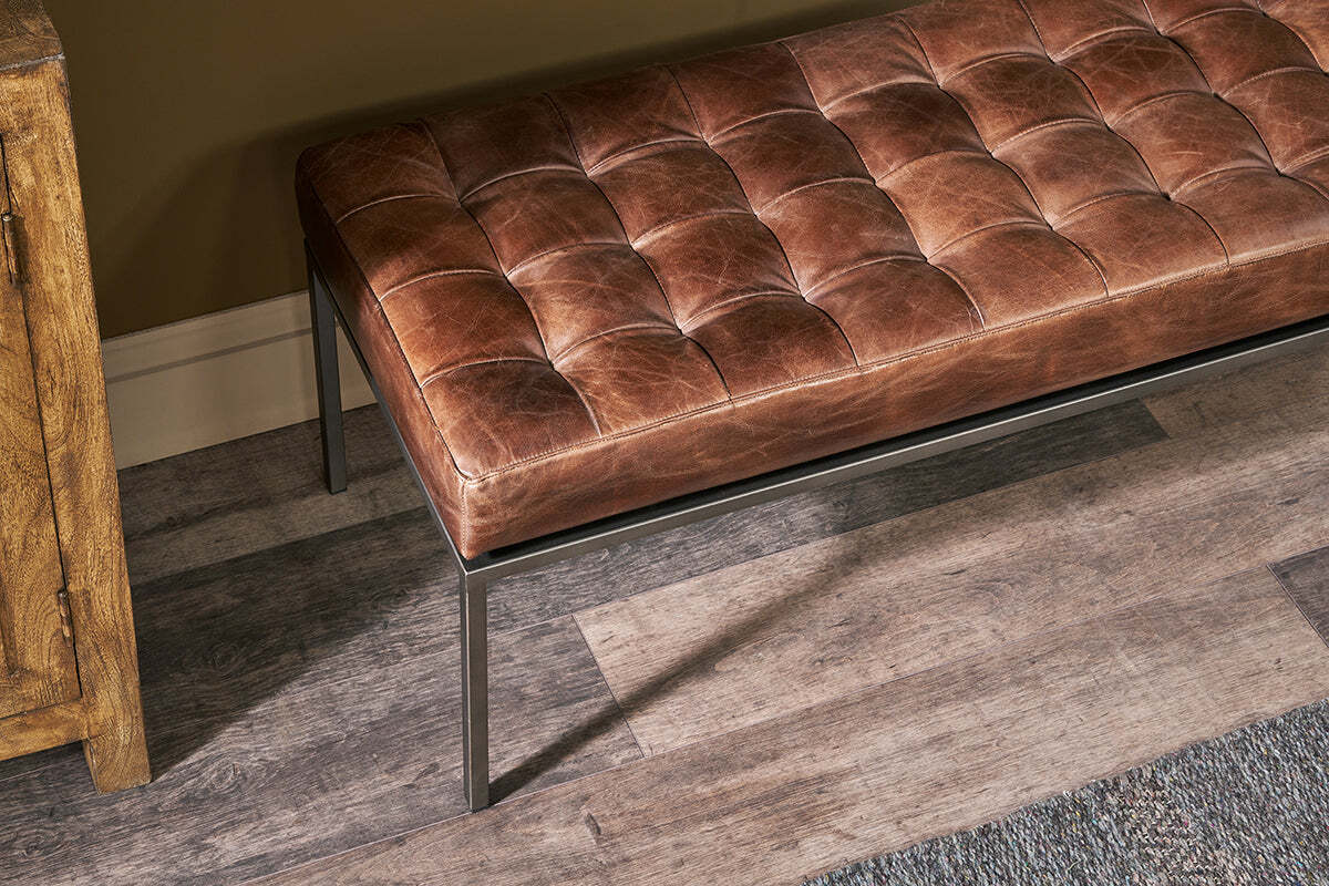 The Nasan Leather Upholstered Bench by nkuku, featuring a tan leather seat and solid iron frame, sits on a wood-look floor beside a wooden cabinet and textured rug, its versatile design enhanced by a tan wall with white baseboard trim.
